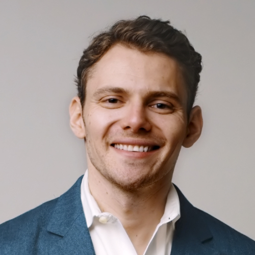 Man with wavy brown hair, white shirt, and gray jacket smiles.